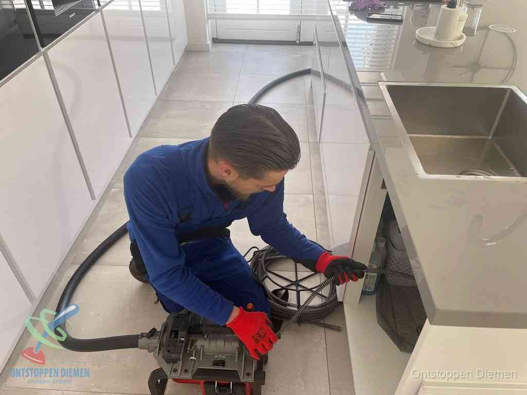 Een monteur ontstopt de keukenafvoer onder de spoelbak met een elektrische veermachine in Diemen