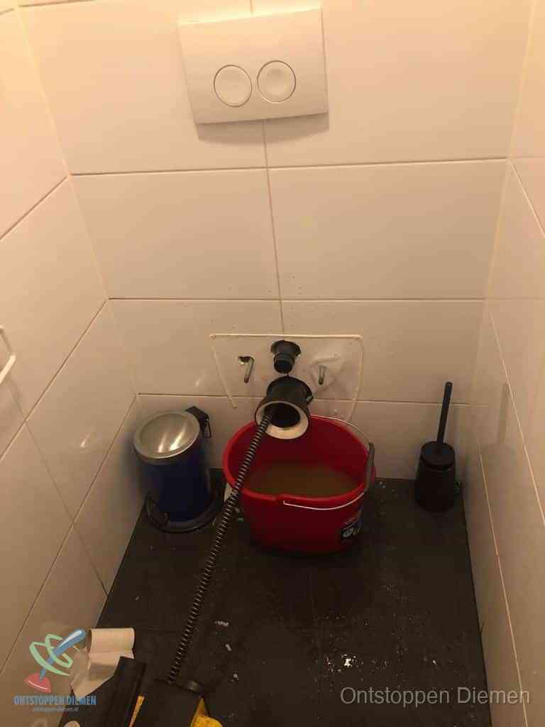 Een ontstopte wand-wc met verwijderde toiletpot waar een ontstoppingsveer in de wanduitgang verdwijnt, het achterhalen waar de verstopping zit in Diemen