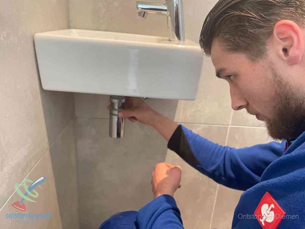Een monteur draait met de hand de zwanenhalssifon onder een fonteinwasbak in de toiletruimte los om de bron van rioollucht in Diemen te vinden