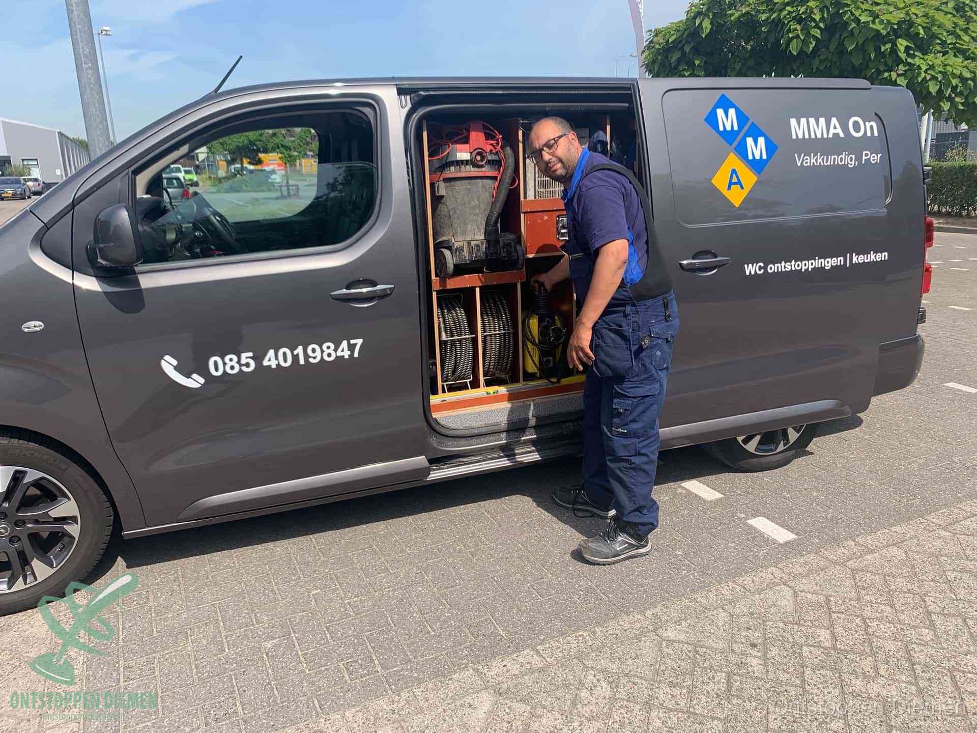 Een monteur bij de open schuifdeur van zijn bedrijfsbus op een parkeerplaats, klaar om de oorzaak van terugkerende afvoerverstoppingen aan te pakken in Diemen