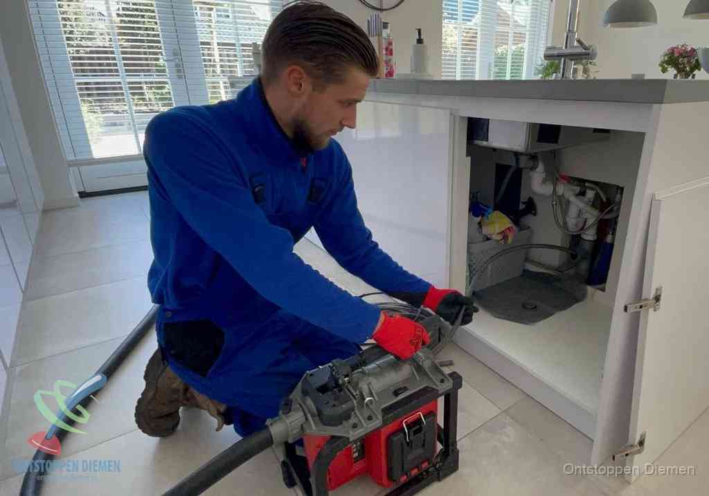 Een monteur voert een professionele elektrische ontstoppingsmachine in de afvoer onder de spoelbak tijdens het gootsteen ontstoppen in Diemen